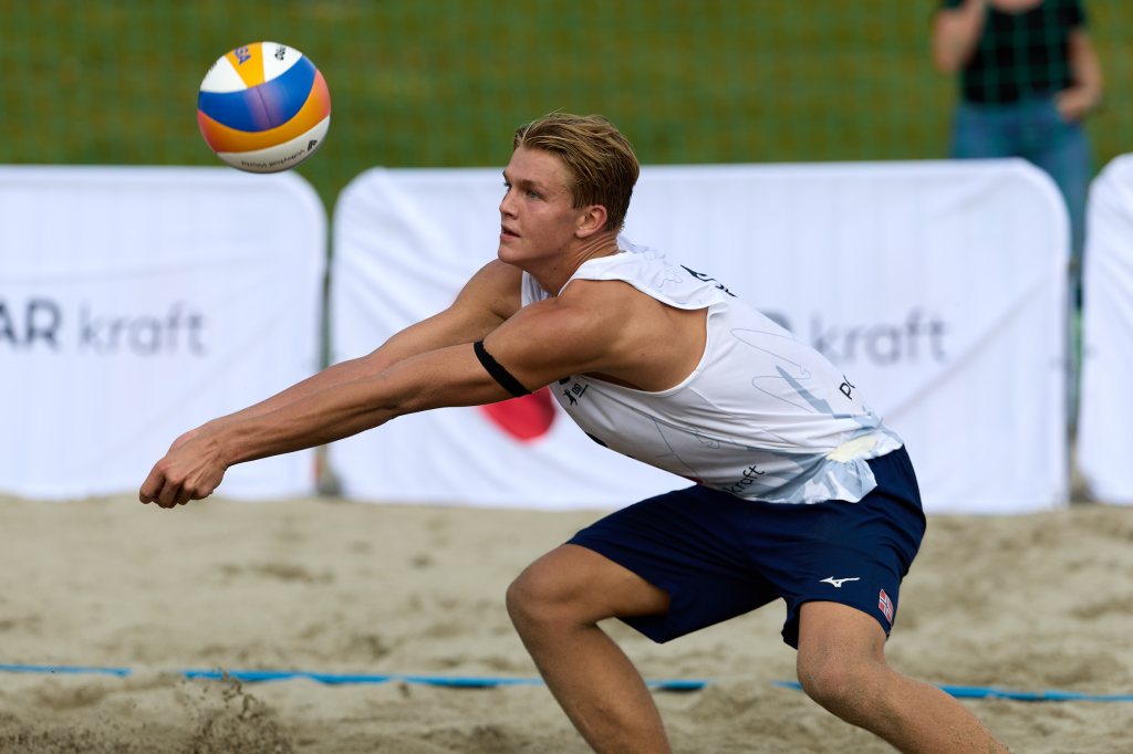 En ung mann i en sportsdrakt spiller sandvolleyball, klar til å mottake en ball som flyr mot ham.