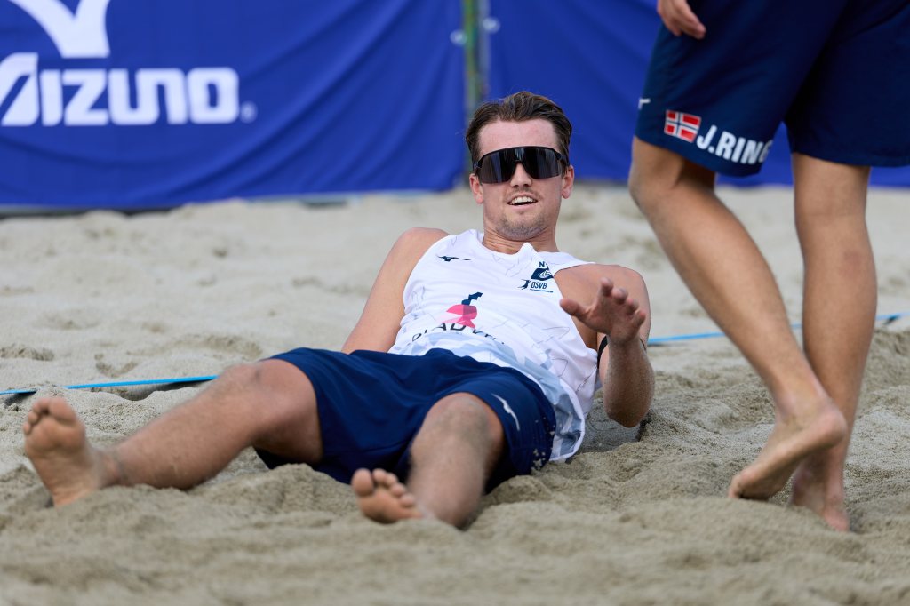 En mann iført solbriller ligger på sanden under en beachvolleyballkamp, med en annen person i nærheten.