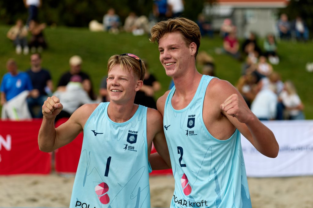 To glade beachvolleyballspillere feirer etter en seier, iført lyseblå drakter. Bak dem er en folkemengde som ser på.