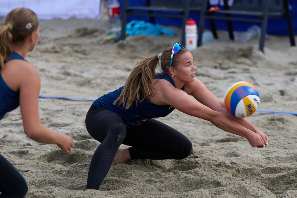 To kvinner spiller beachvolleyball på sand. Den ene spilleren bøyer seg ned for å ta imot ballen.
