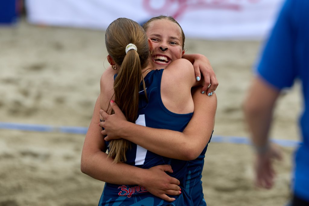 To jenter i sportstøy klemmer hverandre etter en seier på en sandvolleyballbane.