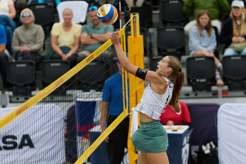 En kvinne som spiller volleyball i sand, hopper for å slå ballen over nettet, med publikum som ser på i bakgrunnen.