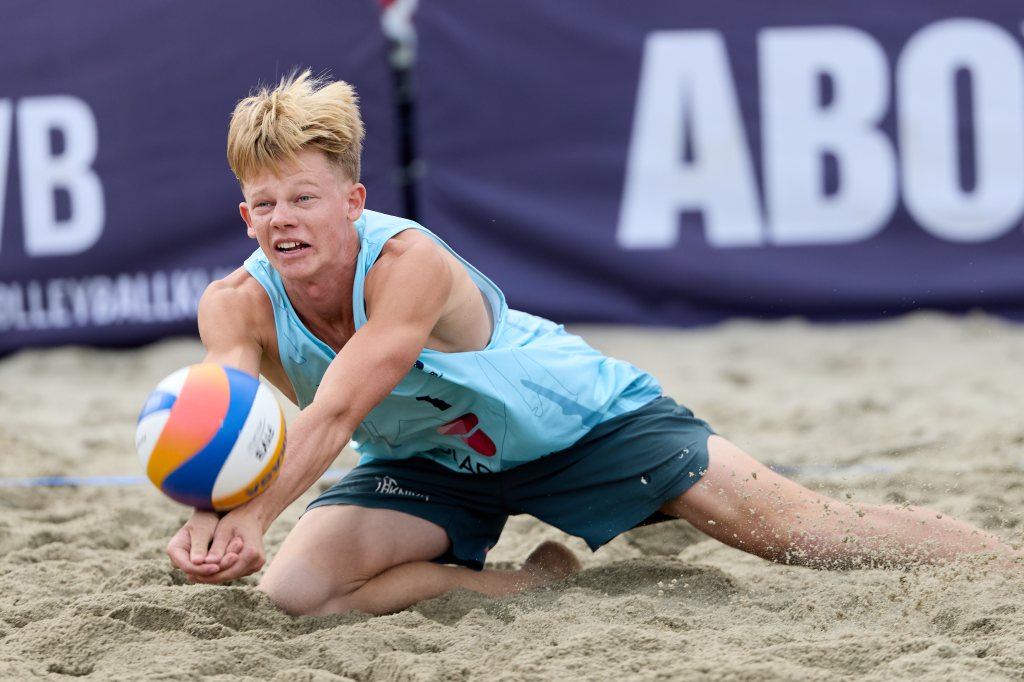 Ung mann spiller sandvolleyball, dypt ned på kne med fokus på ballen, foran en bakgrunn med sponsor-logo.