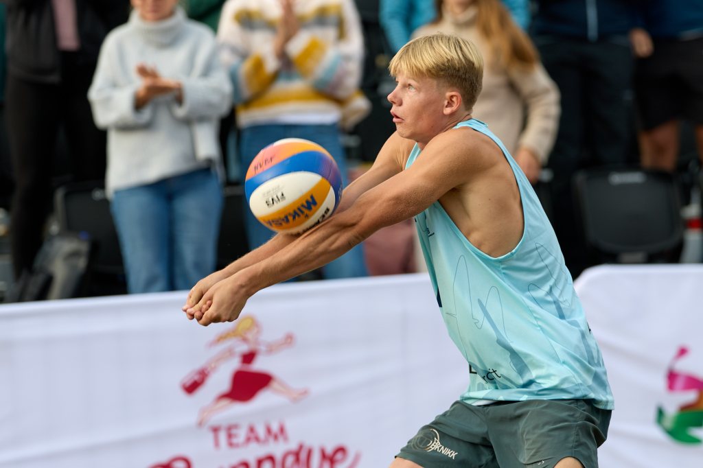 En ung mann i en blå treningstop som spiller volleyball, klar til å ta imot ballen under en konkurranse. Folk klapper i bakgrunnen.