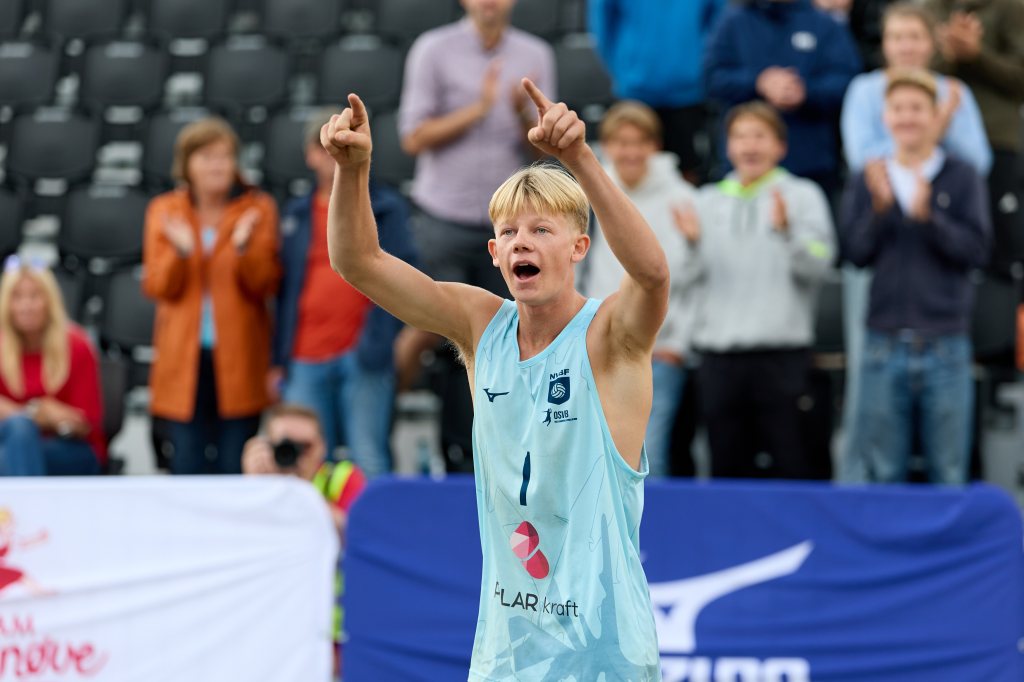 Ung mann i sportsdrakt hever hendene og jubler etter å ha vunnet en konkurranse, med tilskuere i bakgrunnen som applauderer.