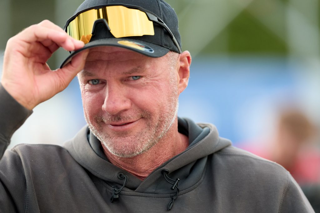 En smilende mann med grå skjegg og blå øyne, iført caps med gullbriller og en grå hettegenser.