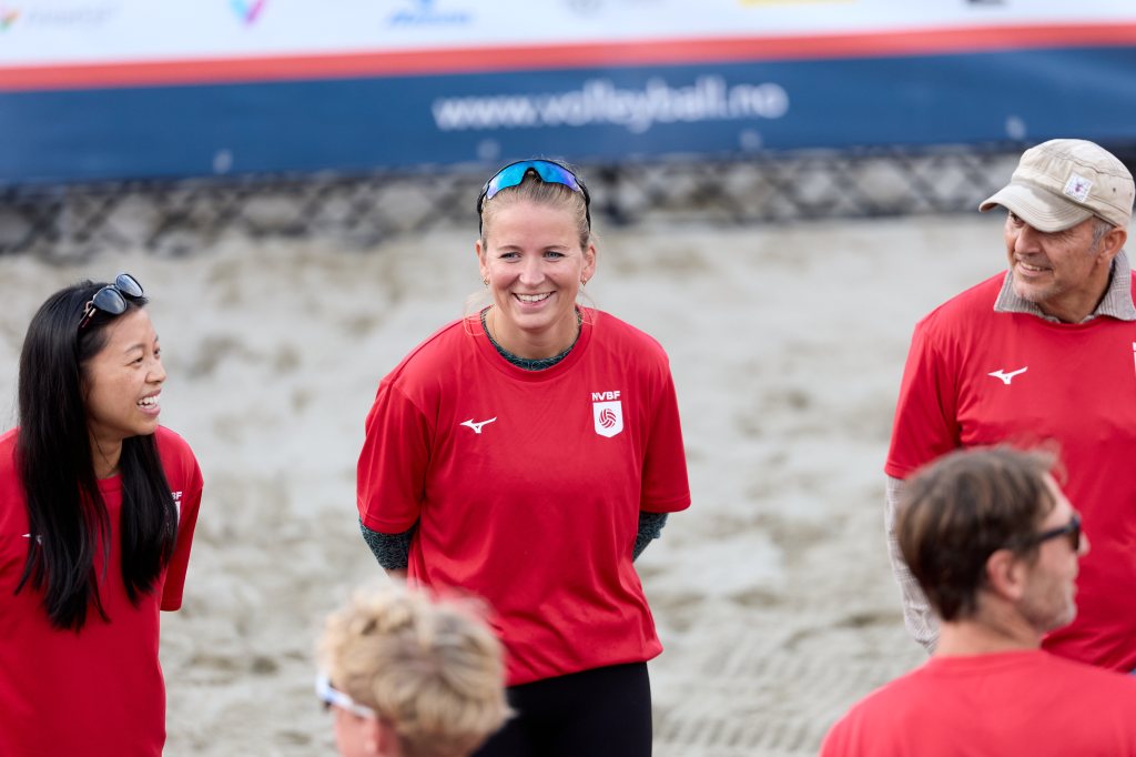 En gruppe mennesker i røde skjorter smiler og snakker sammen på en strand, med sand i bakgrunnen og en volleyballbanner bak.