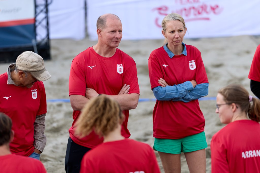 Gruppe mennesker i røde T-skjorter samles på en strand, med fokus på en kvinne og en mann som ser alvorlige ut.