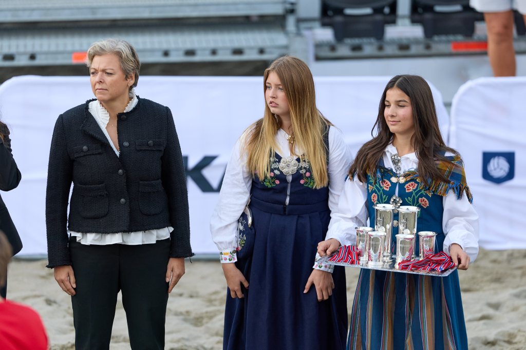 Tre personer står på en strand, med en kvinne iført en svart jakke og to jenter iført tradisjonelle norske drakter. Den ene jenta holder et fat med sølvbeholdere.