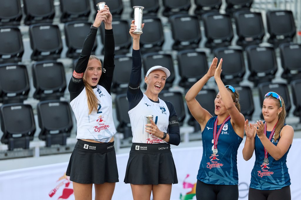 To kvinnelige volleyballspillere hever sine troféer i jubel etter en konkurranse, mens to andre spillere applauderer. Den ene spilleren har på seg en hvit drakt med teksten 'Team Synnyrve', mens den andre har en svart drakt. Alle har medaljer rundt halsen.
