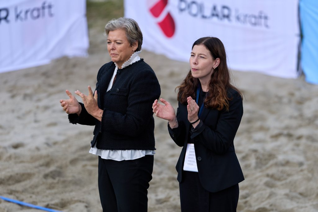 To kvinner klapper under en utendørs begivenhet med sandbakke, iført formelle klær, med sponsorskilt i bakgrunnen.